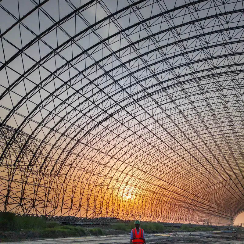 Site engineer in hard hat and hi-vis vest beneath a vast steel space-frame shed at sunset