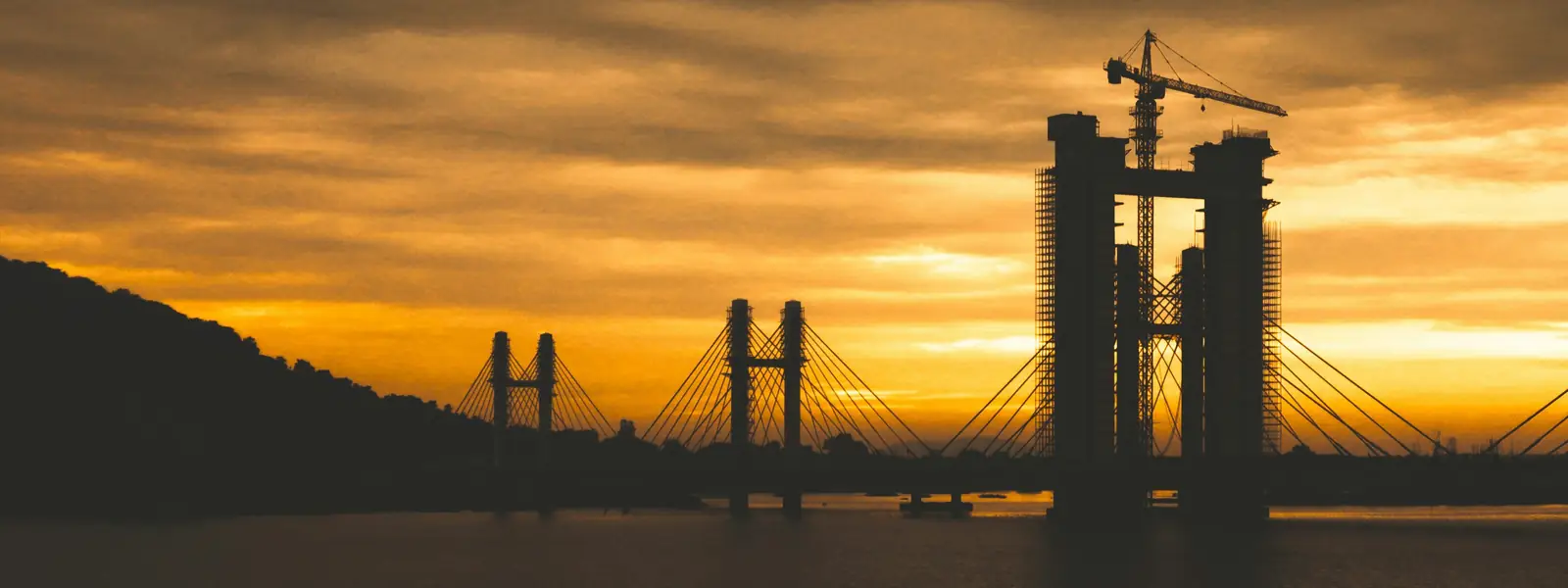 Cable-stayed bridge under construction silhouetted with tower cranes against a dramatic golden sunset sky