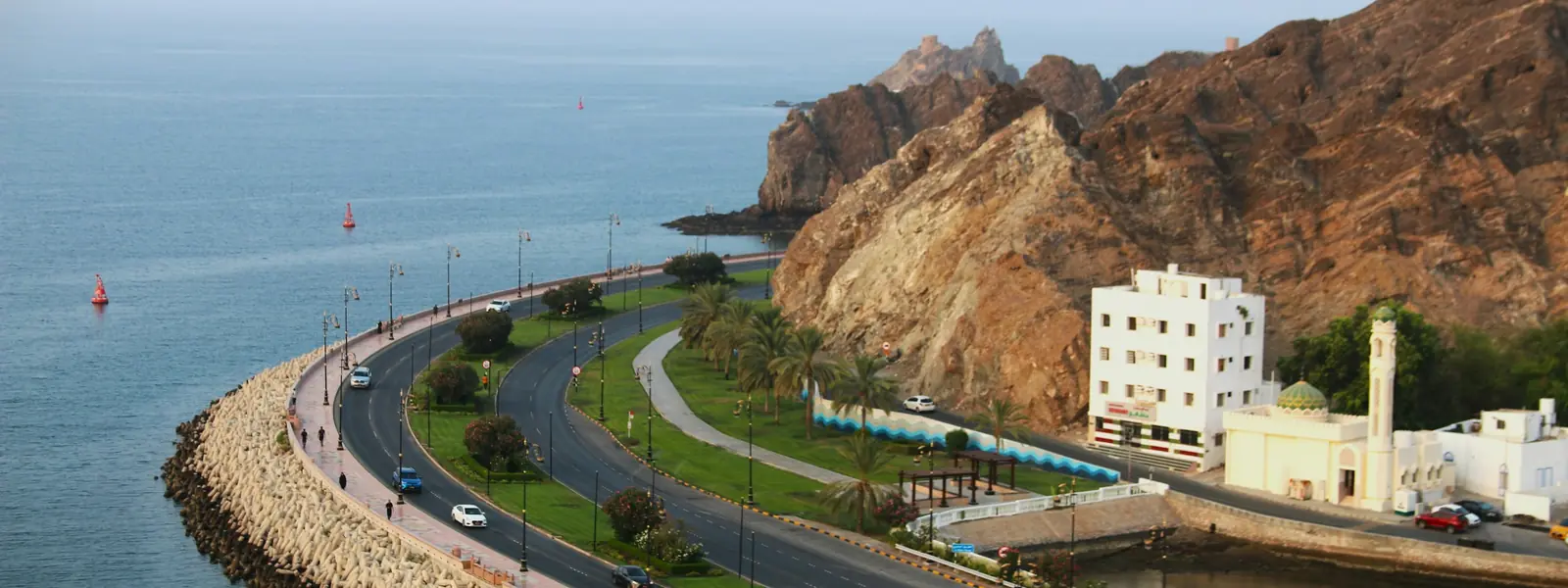 Aerial view of Muttrah Corniche road and breakwater curving along the coast beneath rocky Muscat mountains
