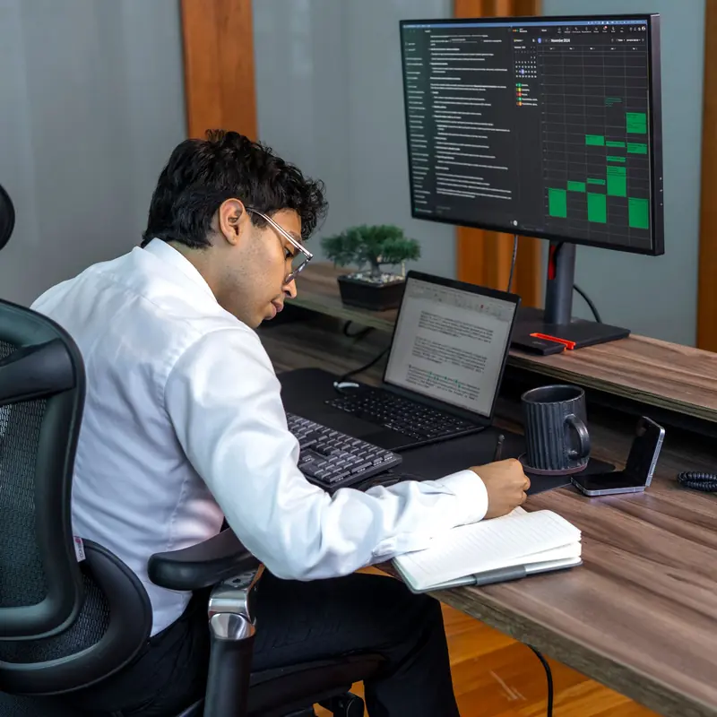 Planner at his desk reviewing a schedule on screen and noting programme actions